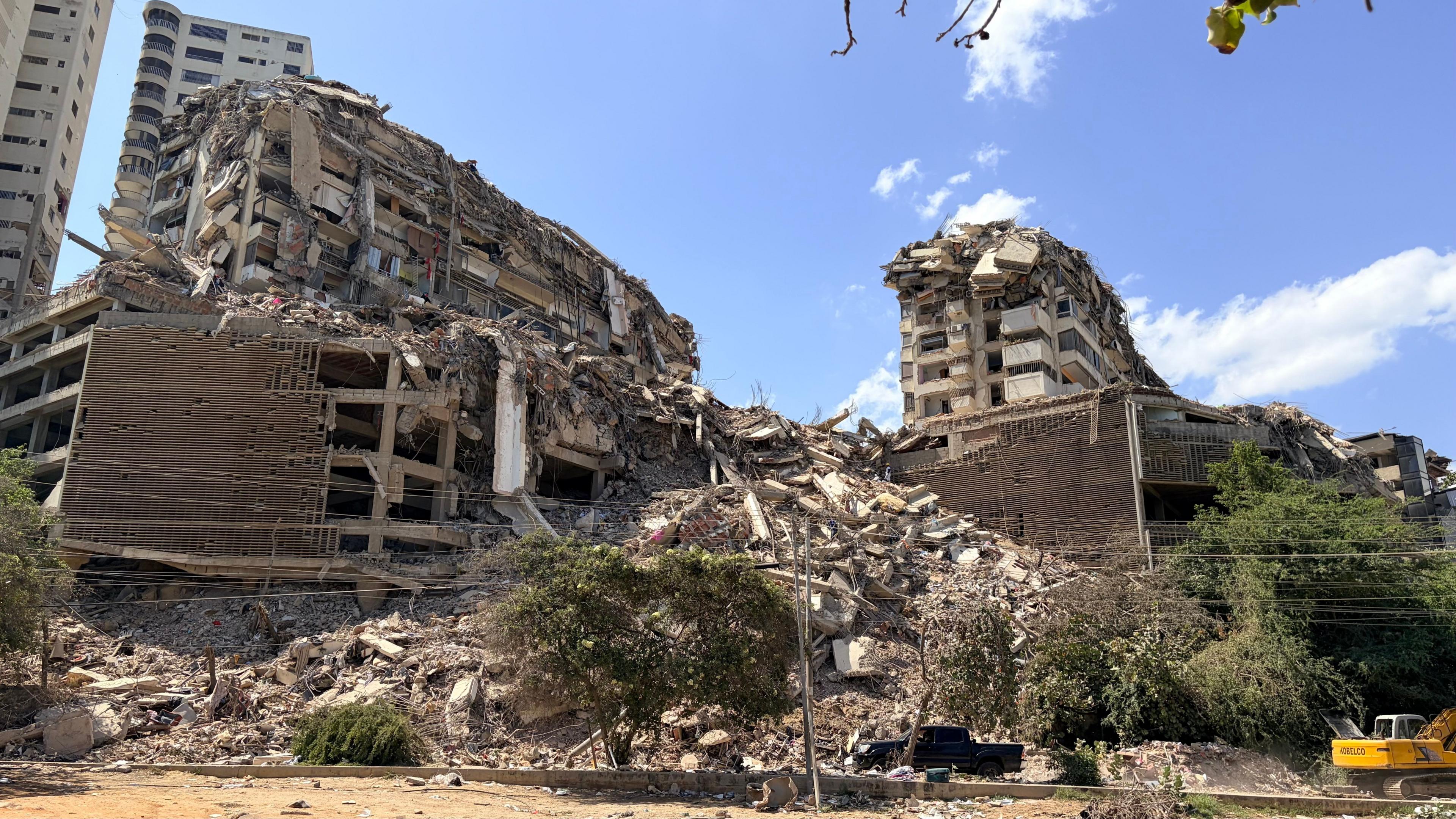 Dramatically collapsed buildings behind a dusty roadside lined with trees