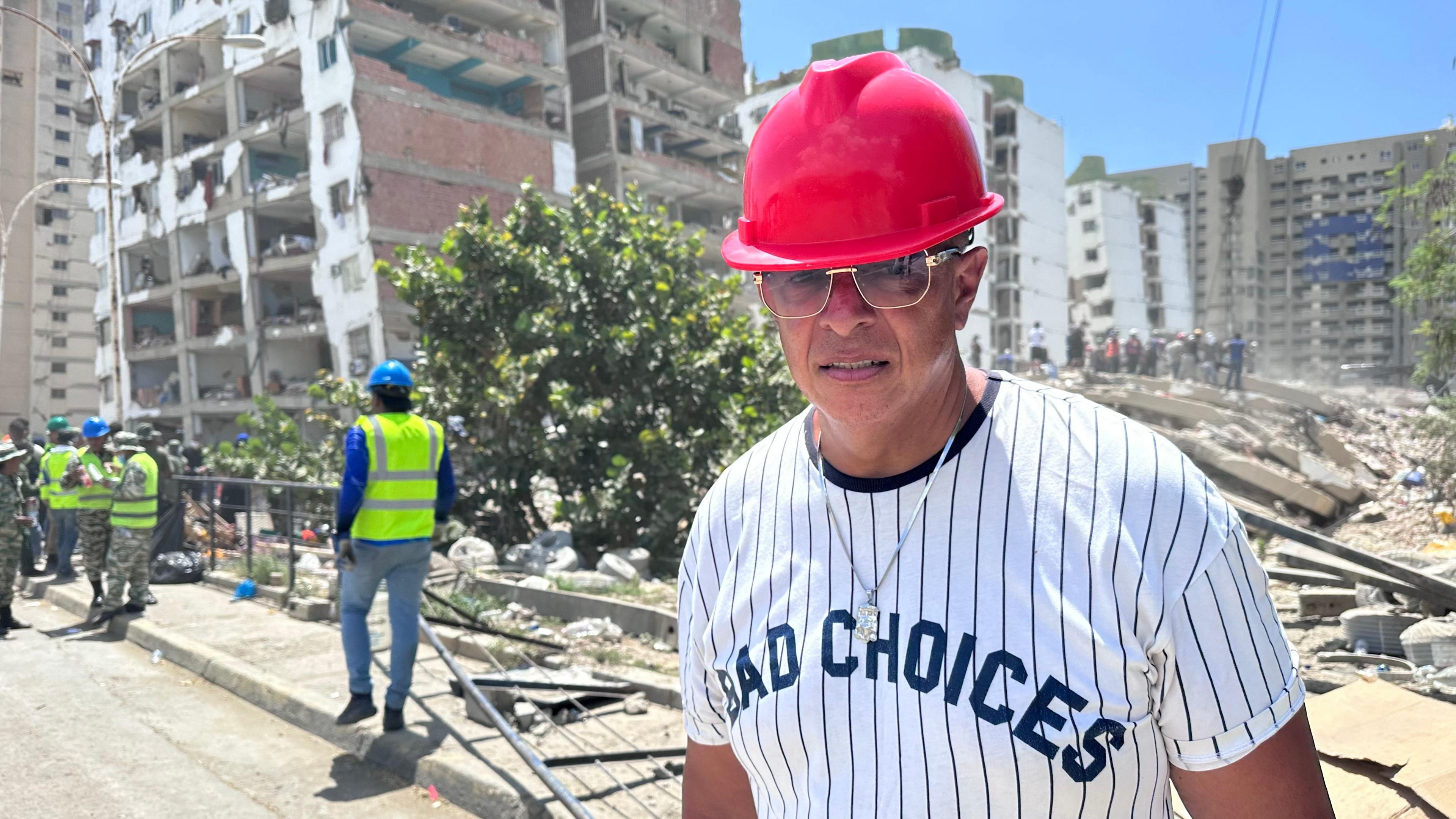 A man in a red hard hat and sunglasses stands on a street in front of the rubble of a collapsed building, with officials in high-visibility vests nearby.