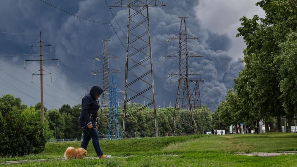 A woman walks in a park as smoke billows in the background following a reported Ukrainian drone attack, in Moscow, Russia, 18 June 2026.