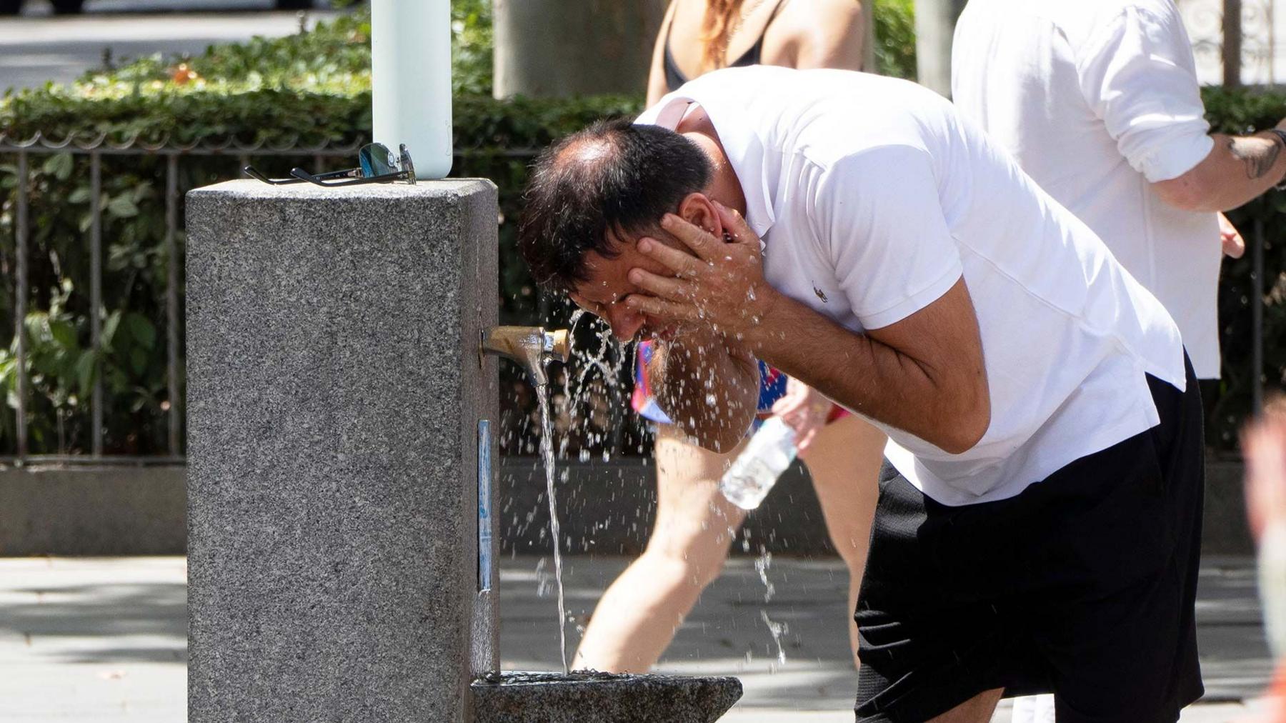 A man splashes his face with water at a drinking fountian in Spain during the heatwave. He is wearing a white T-shirt