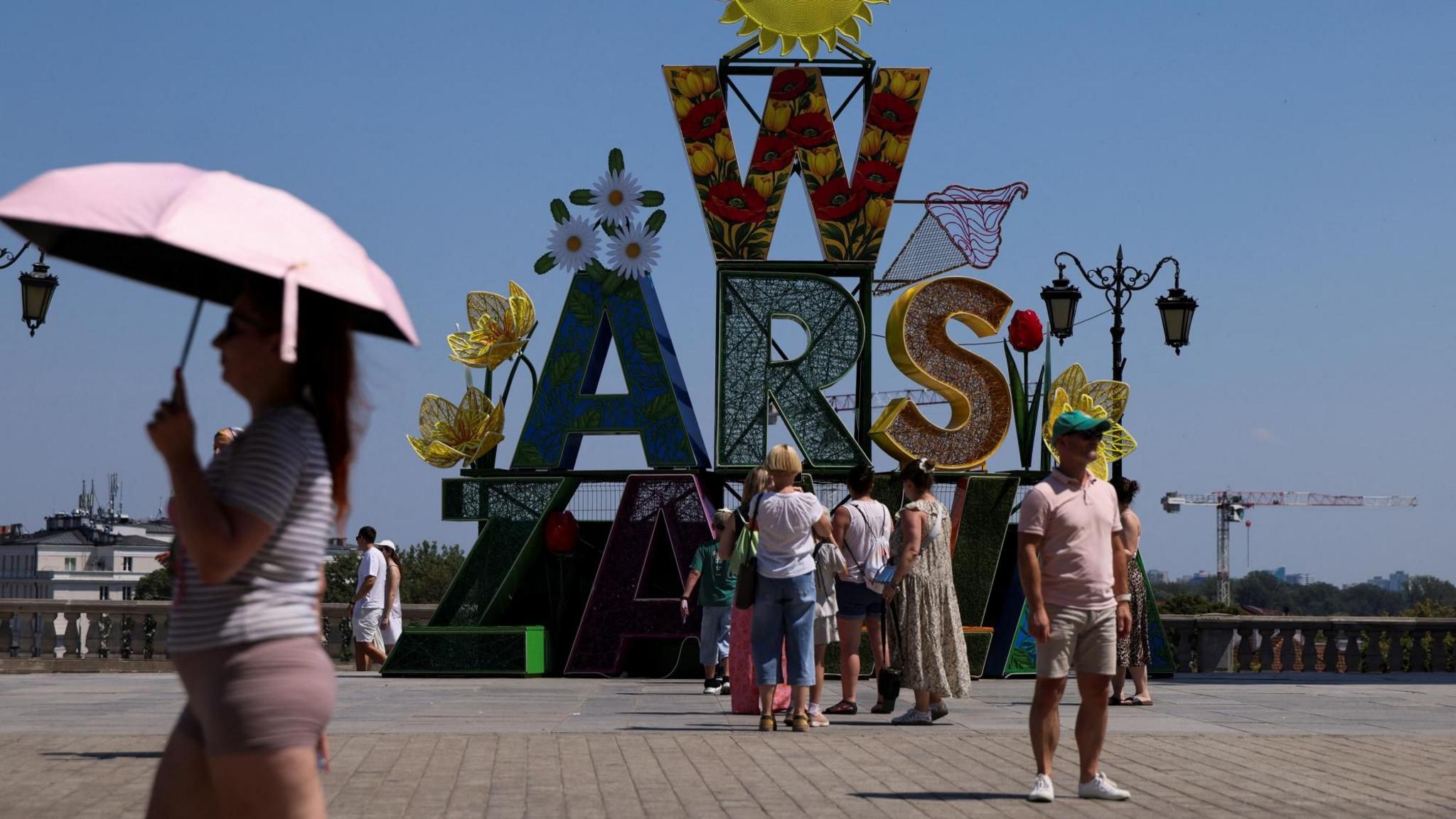 A woman walks under an umbrella past a "Warsaw" sign as a heatwave sweeps Poland, in Warsaw, Poland,
