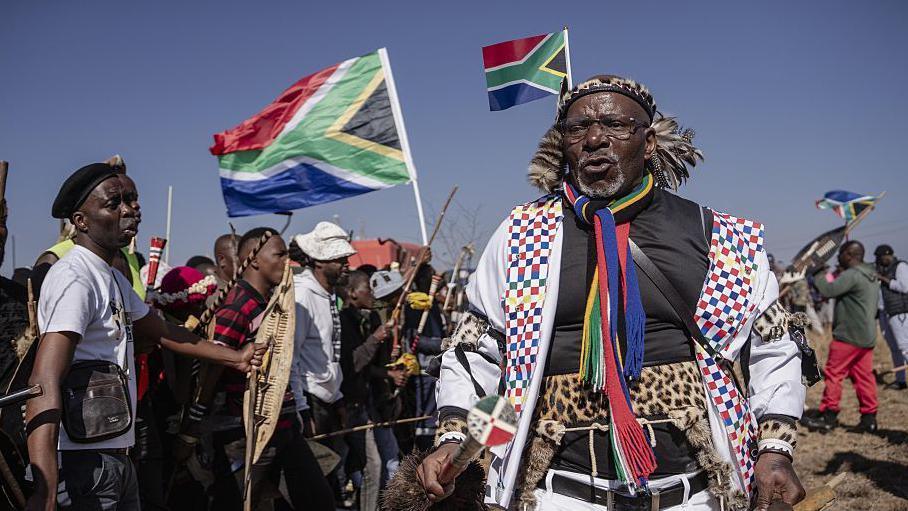 Hundreds of protesters marching outdoors, chanting and holding south african flags and signs