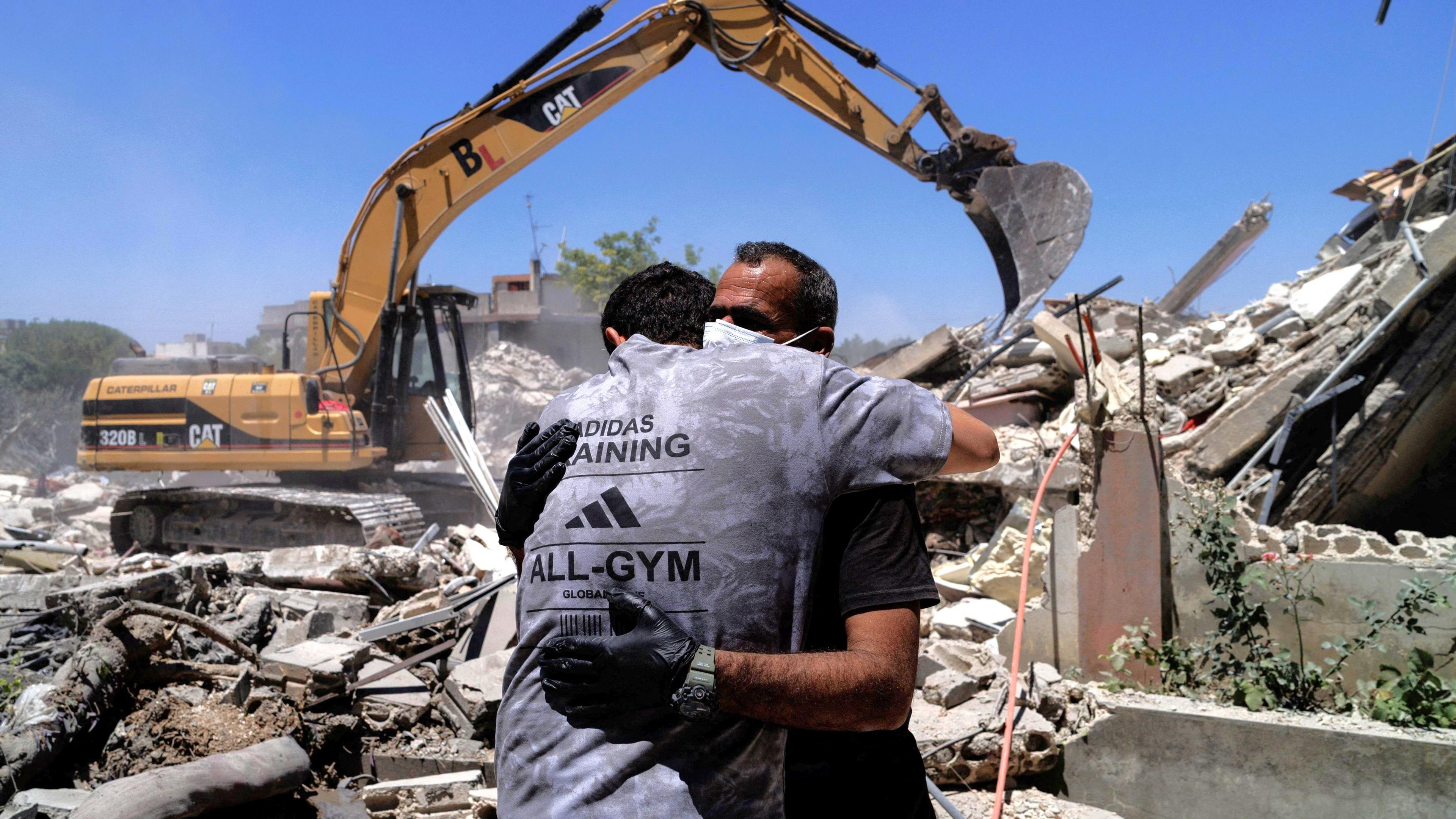 A man wearing a grey Adidas t-shirt hugging another man wearing a black t-shirt, gloves and surgical mask standing in front of a digger clearing up rubble in the aftermath of an Israeli airstrike in southern Lebanon.