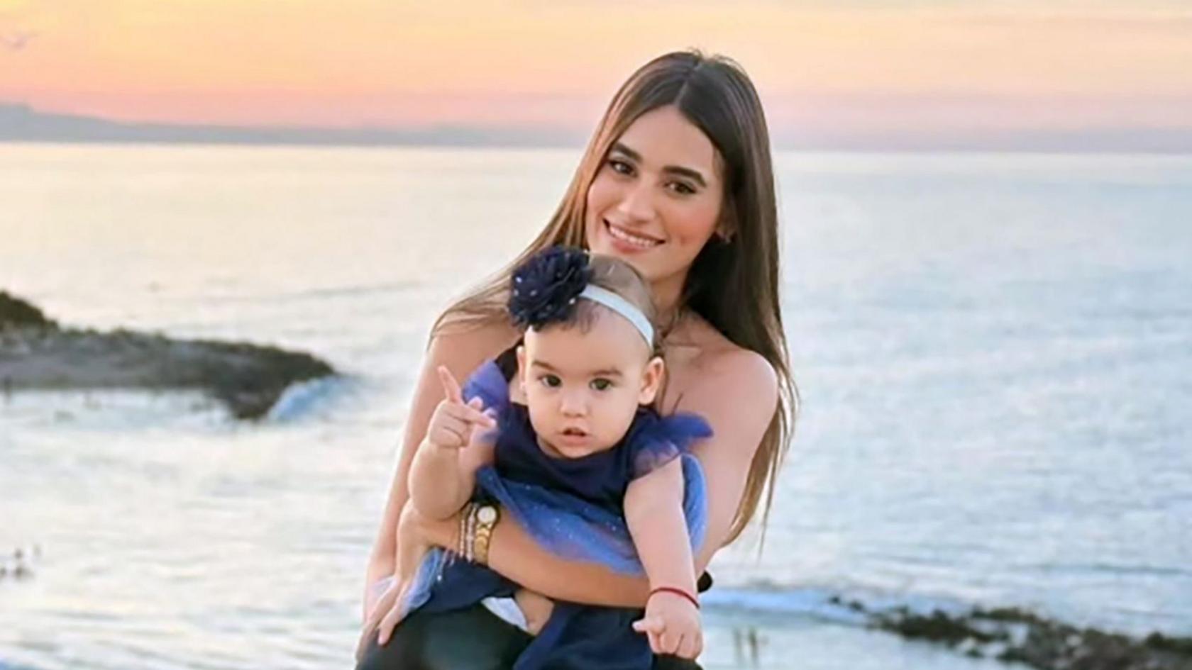 A woman with long dark hair smiles as she holds a toddler in a dress on a beach, the sunset behind her