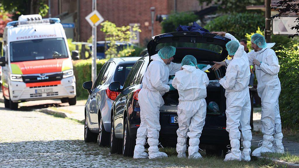 A group of officials in forensic uniforms gathered around the open boot of a black car on the side of a road in Germany.