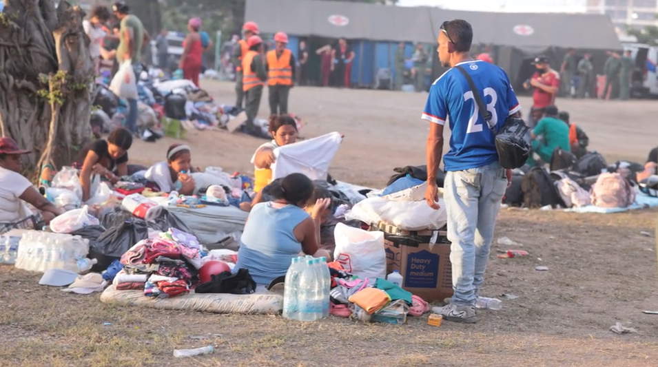 People sit on the ground, surrounded by their possessions, with an emergency tent in the background.