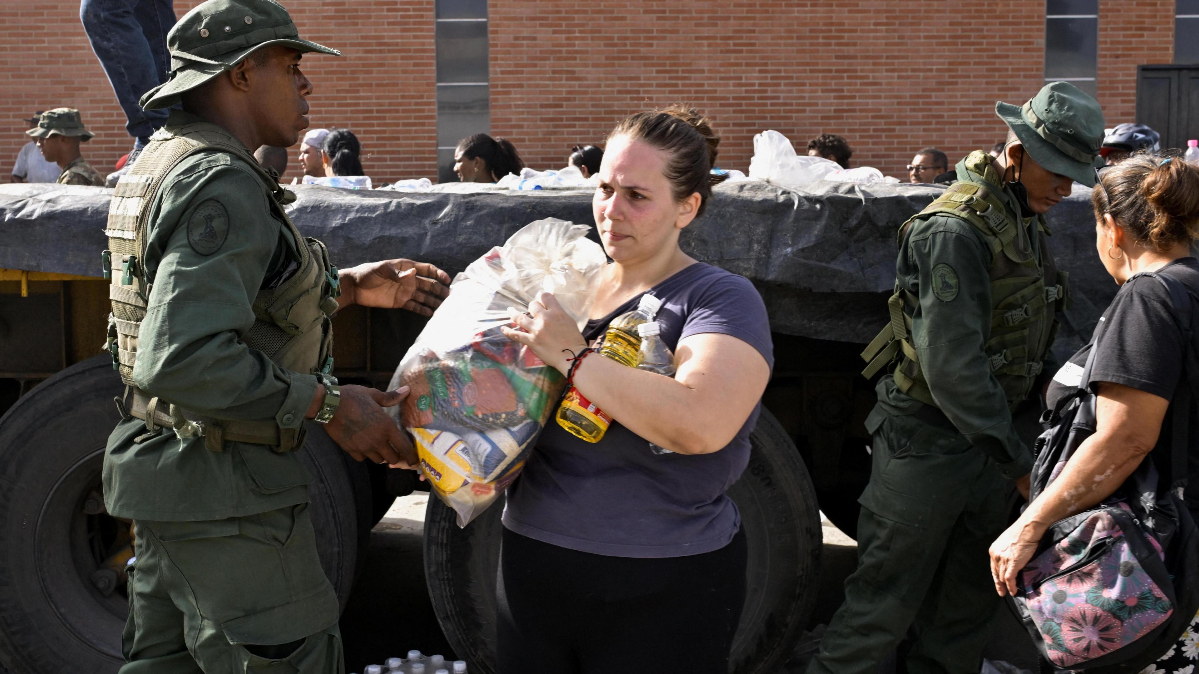 Venezuelan military personnel distribute aid to people affected by deadly earthquakes that hit the country. In the middle of the image, a woman carries a package of food over her shoulder.