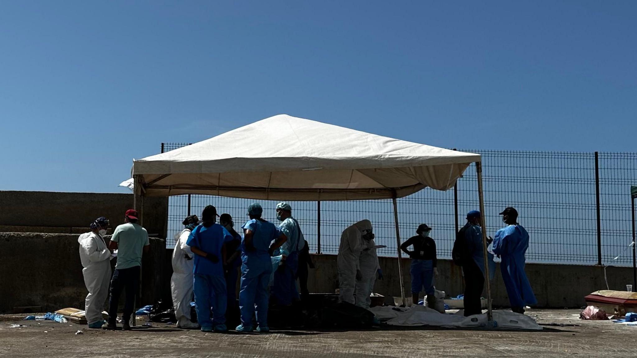 Medical personnel around bodies on the ground covered with plastic bags. The workers are wearing blue or white gowns.