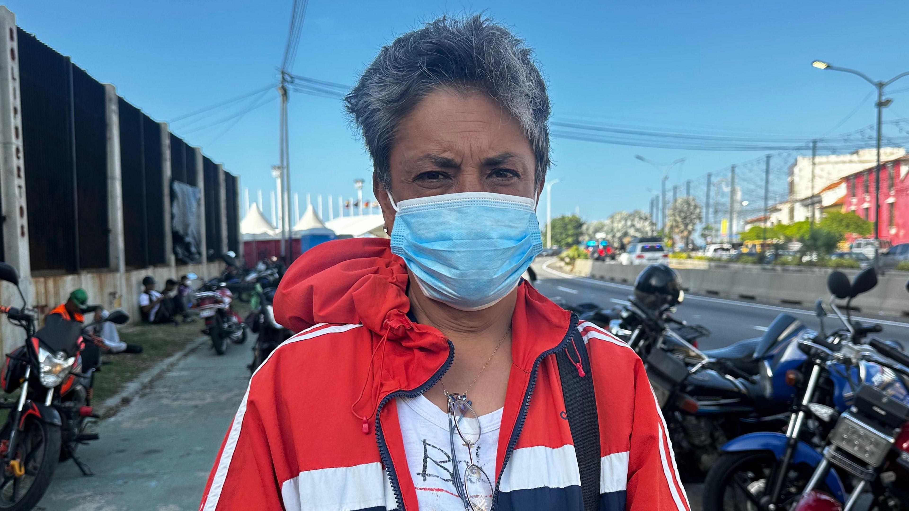 Modesta Alemán, wearing a blue face mask, stands in front of Los Silos. She is wearing a red jacket with blue and white stripes.