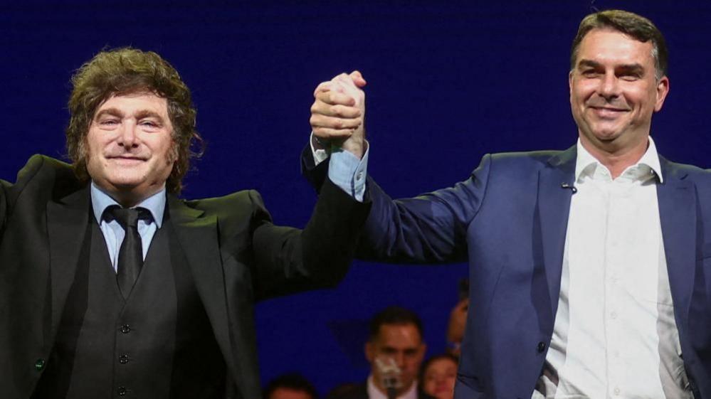 Argentine President Javier Milei holds hands aloft with Brazilian Senator Flavio Bolsonaro. Both men are wearing suits and smiling as they appear at a campaign event.