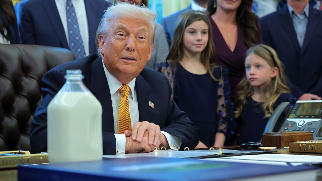 Trump, in a blue suit and yellow tie, sits at his desk in the Oval Office. There are people standing around hi, including two young girls to his left. On his desk is a large bottle filled with milk.