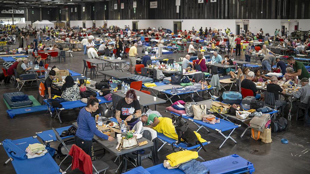 People sit at makeshift table and chairs in a convention centre with supplies on a table