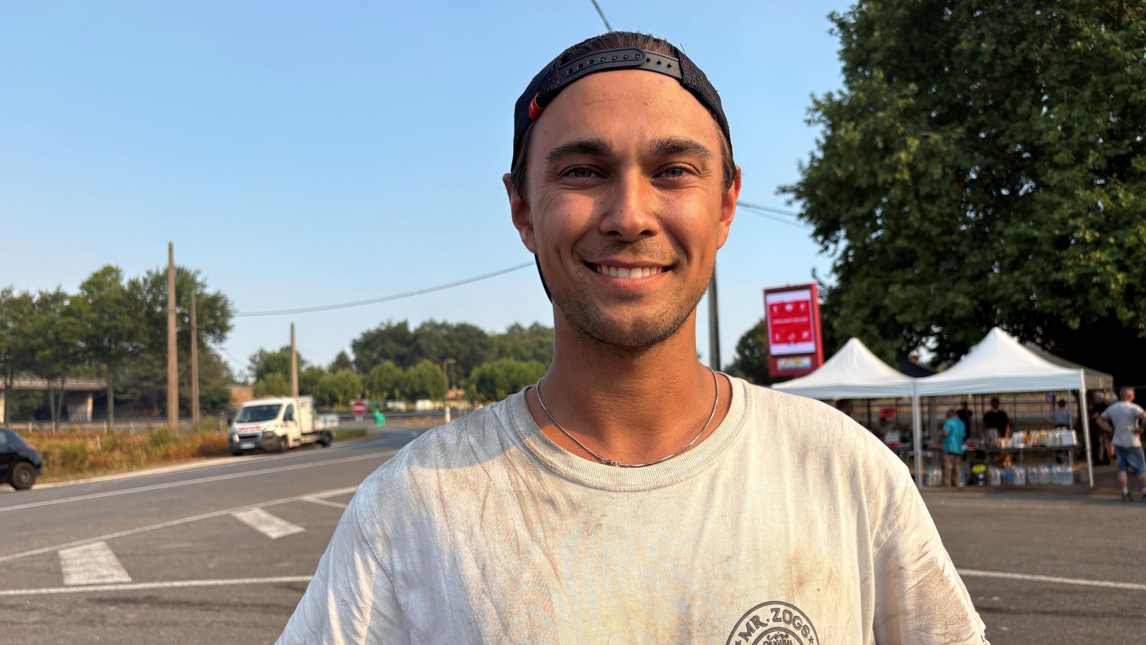 A man wearing a black baseball cap backwards on his head and a white t-shirt grins at the camera as he stands near a road