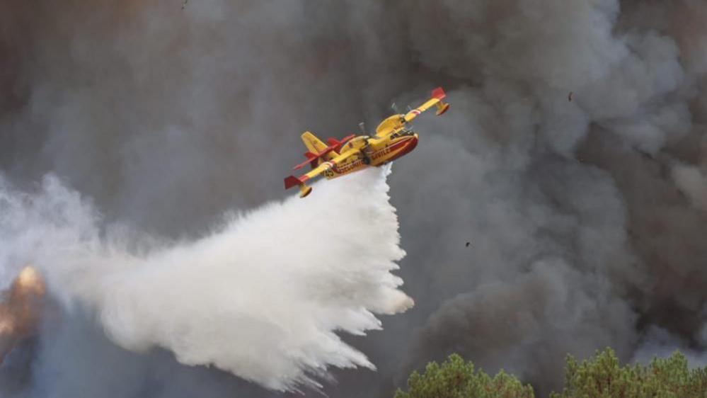 A yellow and red propeller aircraft lists to the left as it drops huge amounts of water. In the ground are enormous black clouds of smoke.