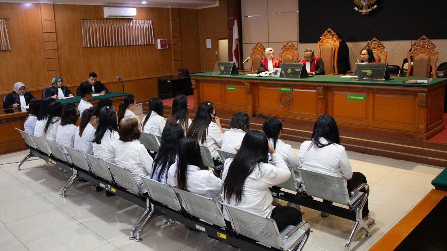 Two rows of women wearing white tops and black trousers sit in front of a judge in a courtroom