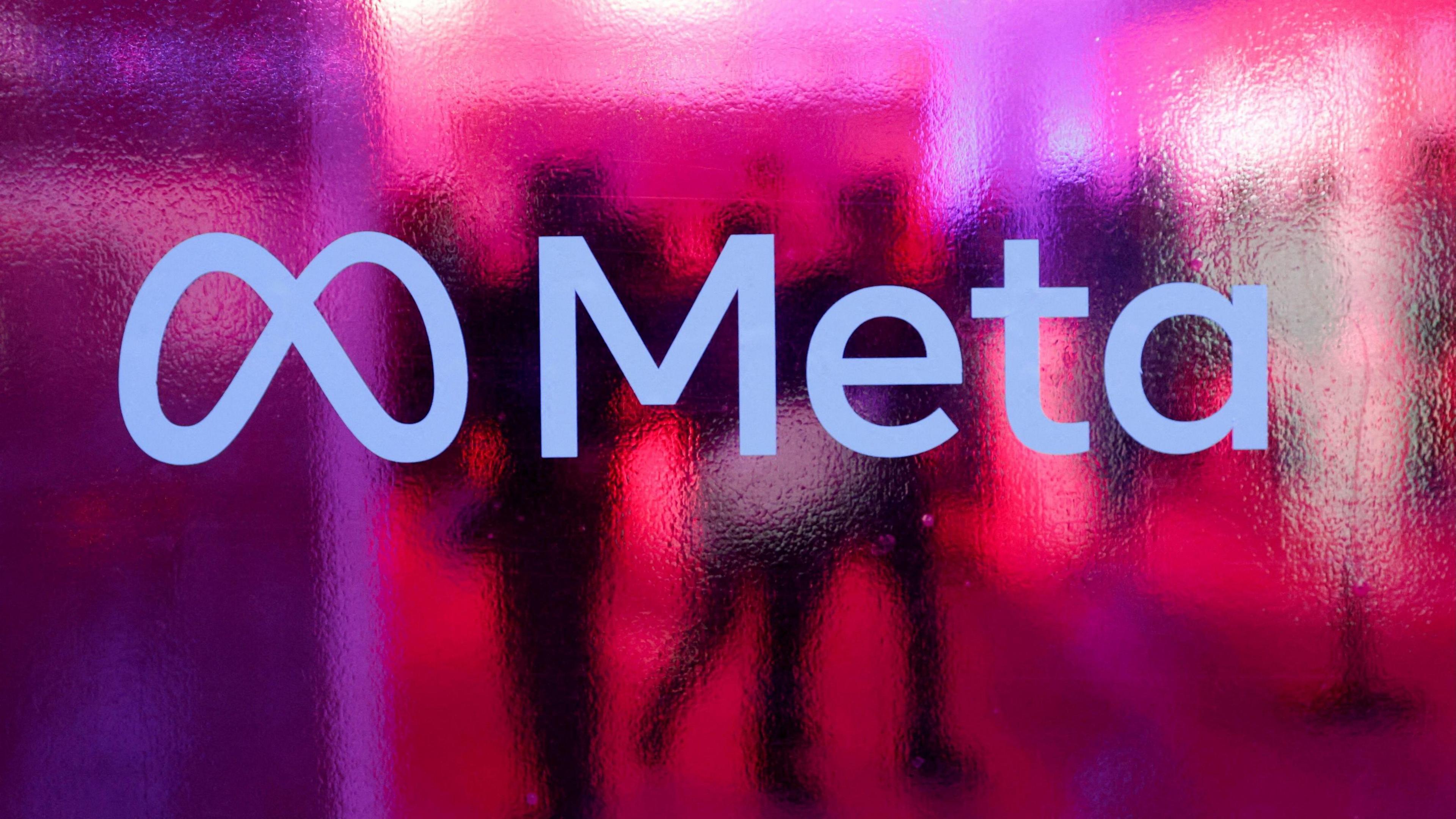 Meta's logo is displayed against pink, frosted glass. Behind the logo are the silhouettes of people.