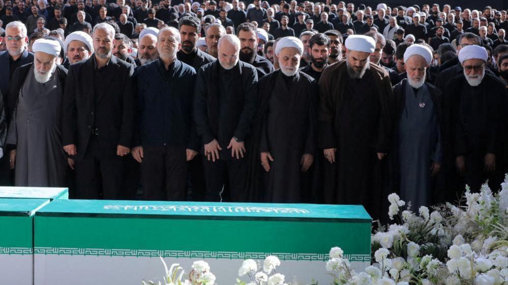 Iranian President Masoud Pezeshkian, Iran's Parliament speaker Mohammad Baqer Qalibaf and Chief Justice of Iran Gholam-hossein Mohseni Ejei with officials attend a prayer during a public farewell ceremony to pay their respects to late Supreme Leader Ayatollah Ali Khamenei. A large crowd can be seen in the background as everyone stands looking at the coffin
