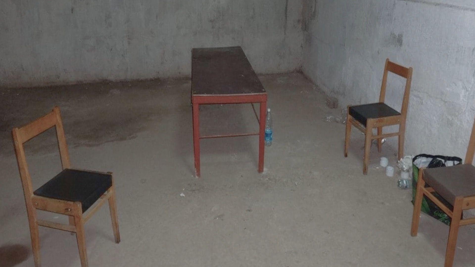 A bare room with concrete walls and floor. In it are three wooden chairs and a small wooden table, with a few plastic water bottles on the floor.