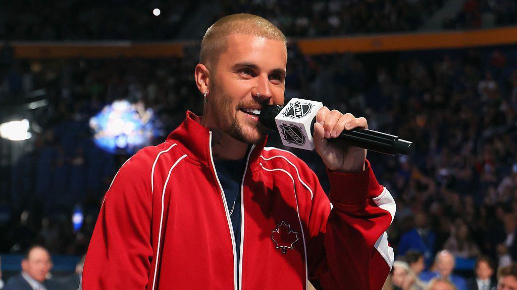 Justin Bieber wears a read jacket and holds microphone with an NHL logo on it while he attends the 2026 NHL Draft on 26 June 2026 in Buffalo, New York