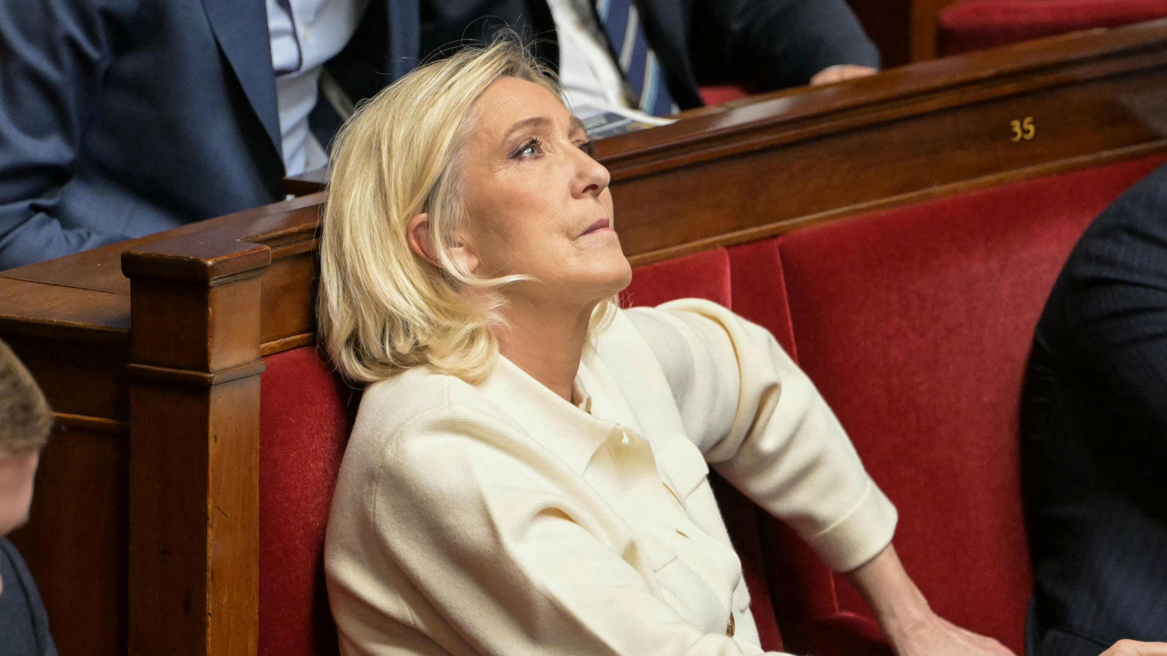 Marine Le Pen sits in France's National Assembly in a red velvet seat. She is biting her lip and looking up. She wears a cream suit.