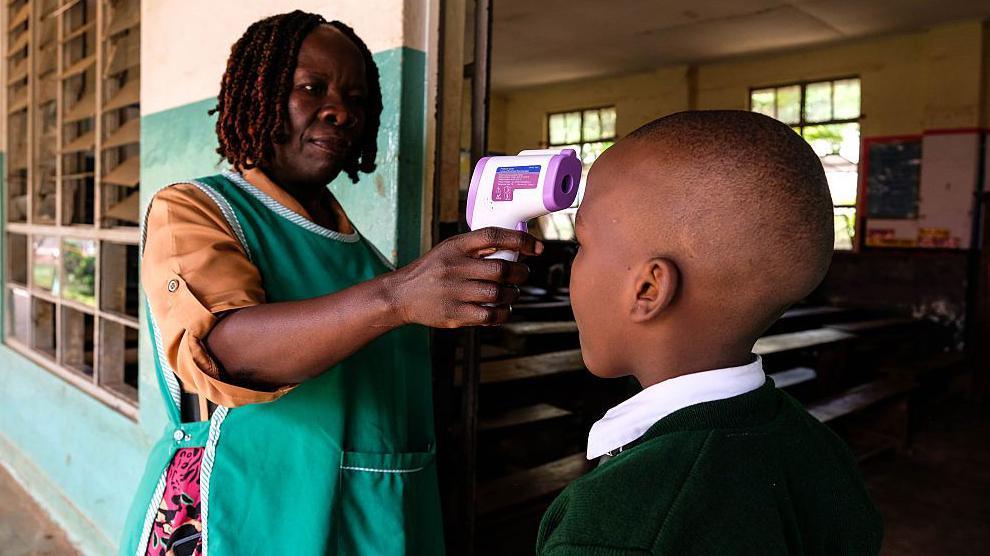A woman in a green tabard uses a thermometer gun to check the temperature of a school pupil who has their back to the camera.