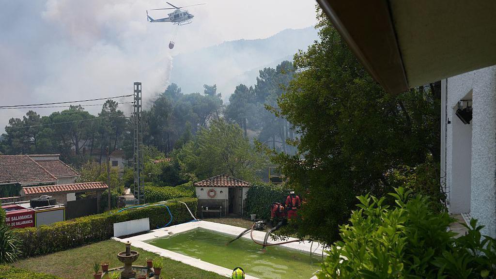Firefighters pump water from a pool with a helicopter dumping water on a forest area filled with smoke