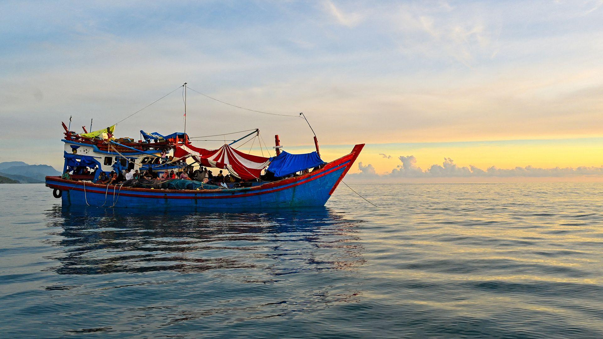 A blue and red boat sailing on the ocean with a sunset in the background with the coast of Indonesia just in view in the left of frame, taken off the coast of Labuhan Haji in Southern Aceh province on October 23, 2024.