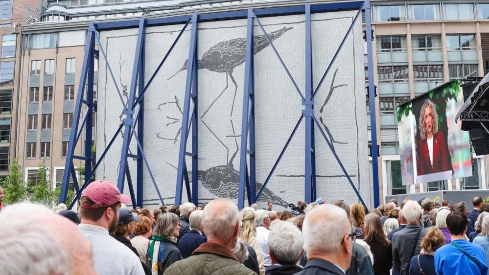 A large crowd stand in front of an artwork depicting a bird reflected in water. The large artwork is covered by a blue steel frame.