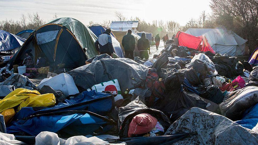 A makeshift camp of tents and tarpaulins stands in an open area during daylight, with piles of rubbish and discarded belongings in the foreground. Several people walk between the tents along a muddy path, and low sunlight casts long shadows across the scene.
