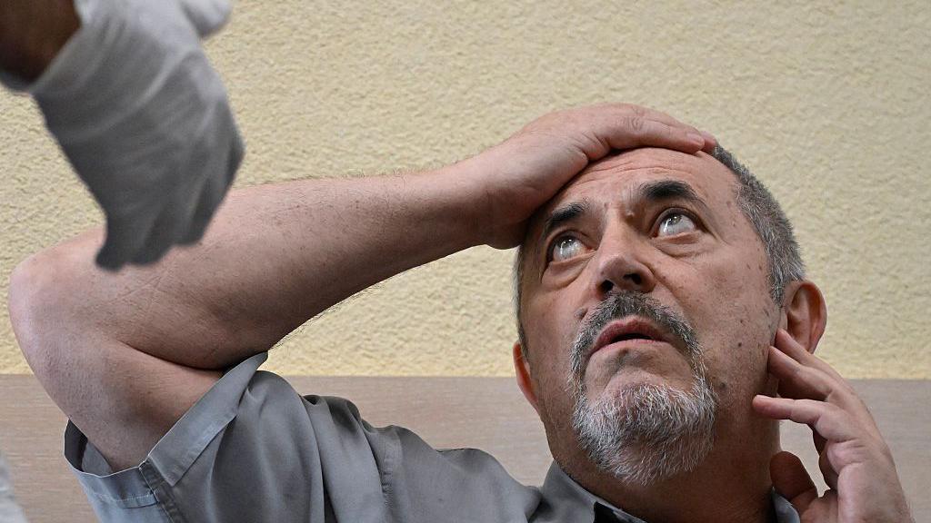 A man in a grey shirt holds his right arm to his forehead as he looks at a man out of the picture who is wearing a medical glove