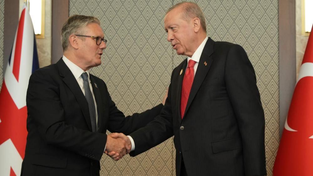 Sir Keir and Erdogan, both wearing dark suits, shake hands. Behind them are the Union Jack and Turkish national flag.