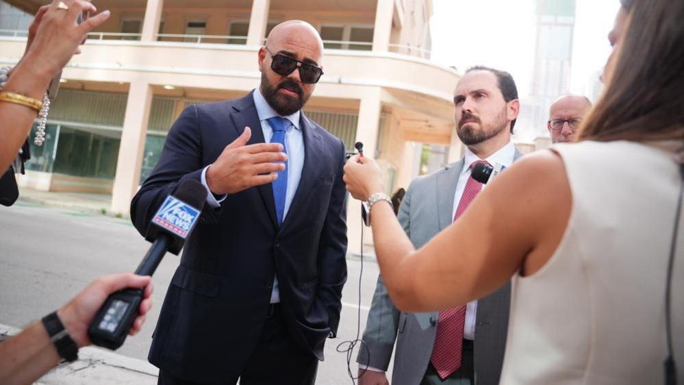 Joseph McBride, bald and with a beard and mustache and wearing sunglasses and a blue suit, shirt and tie, and Thomas Maniotis, with short cropped brown haior and a beard and mustache, with a grey suit and red tie, speak into microphones surrounded by media