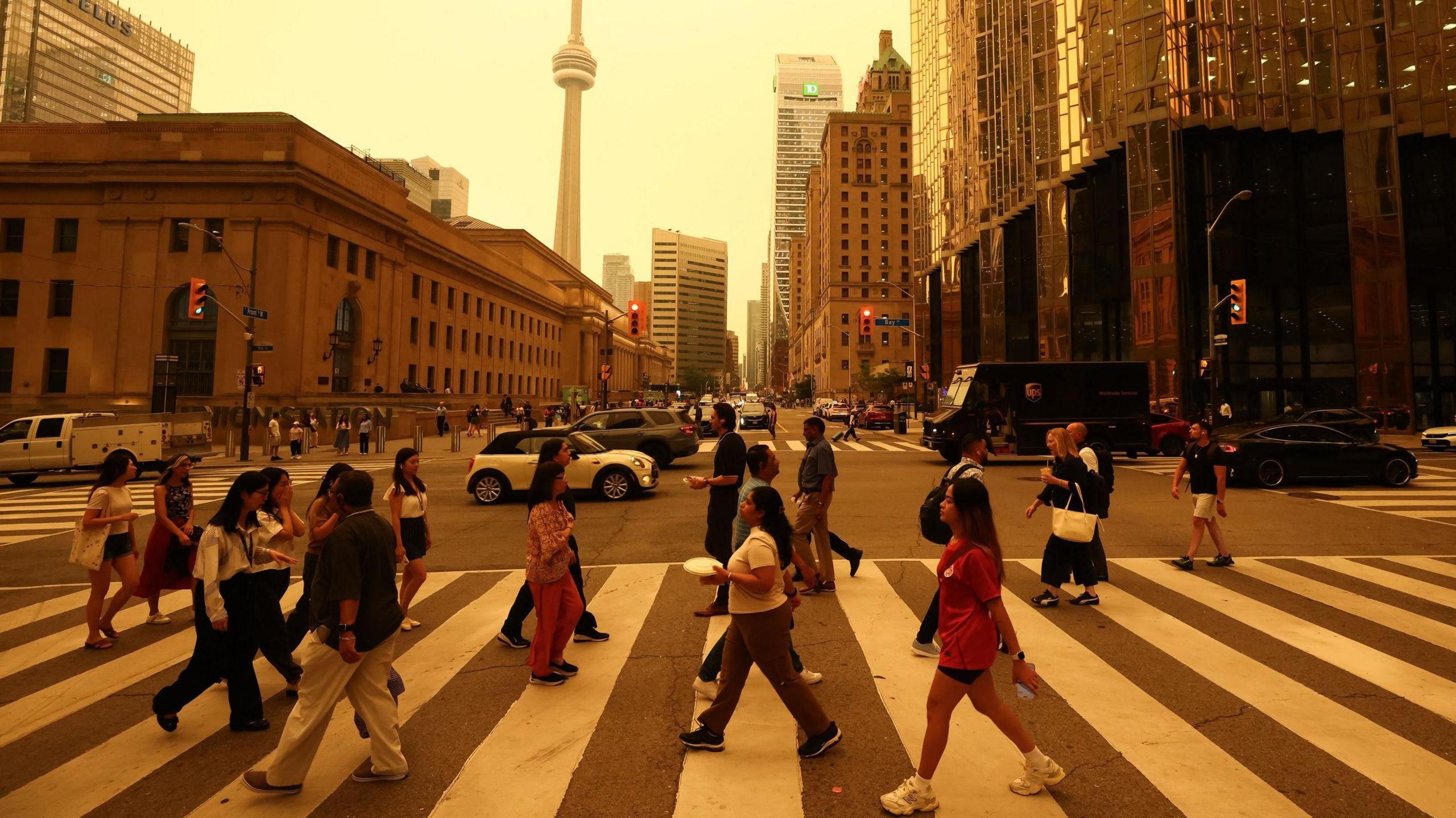 People walk on a crossing in a road downtown Toronto. There are buildings in the background and sky is orange from smoke.