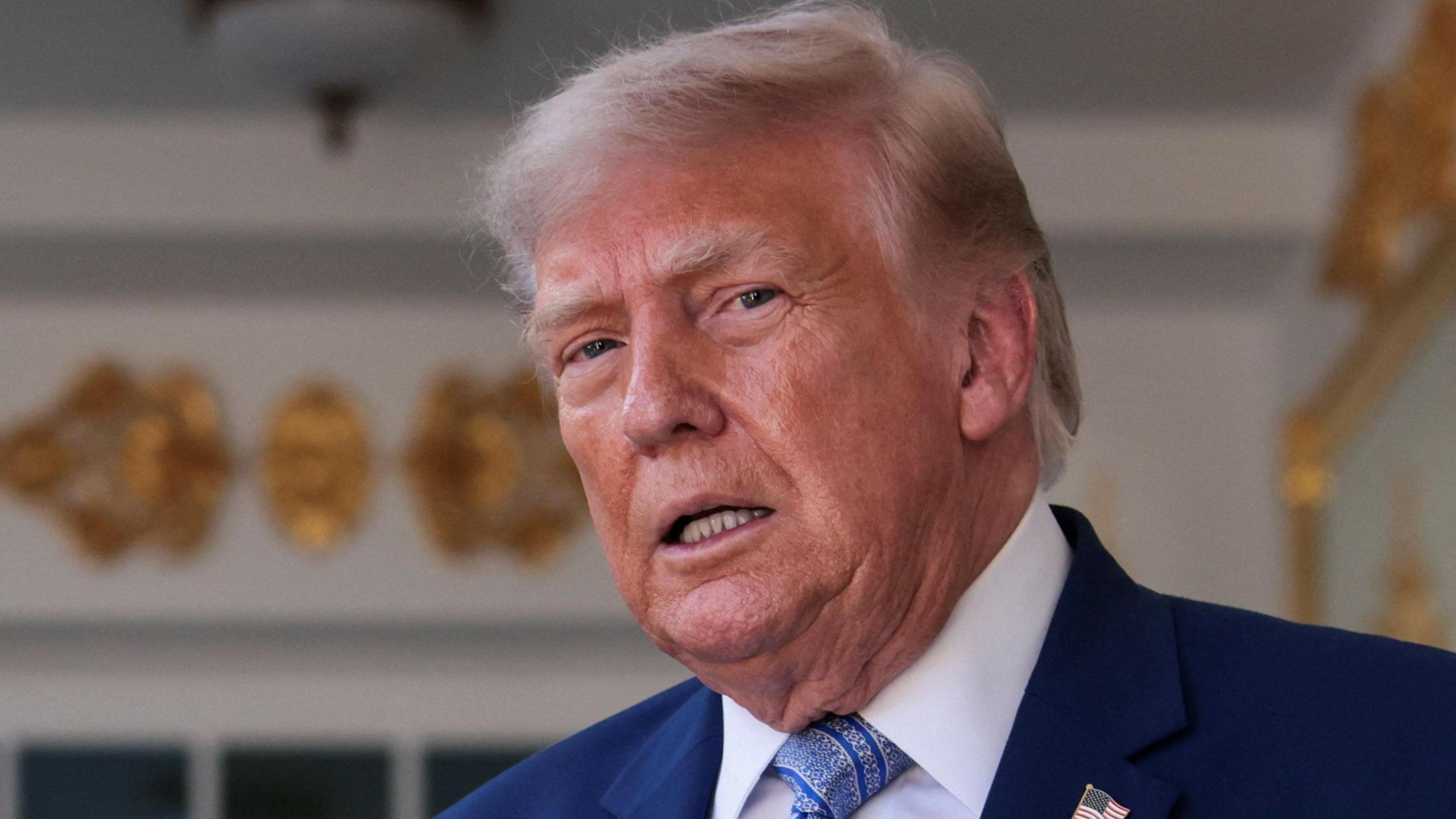 US President Donald Trump speaking in front of a White House entrance, wearing a blue suit and tie.