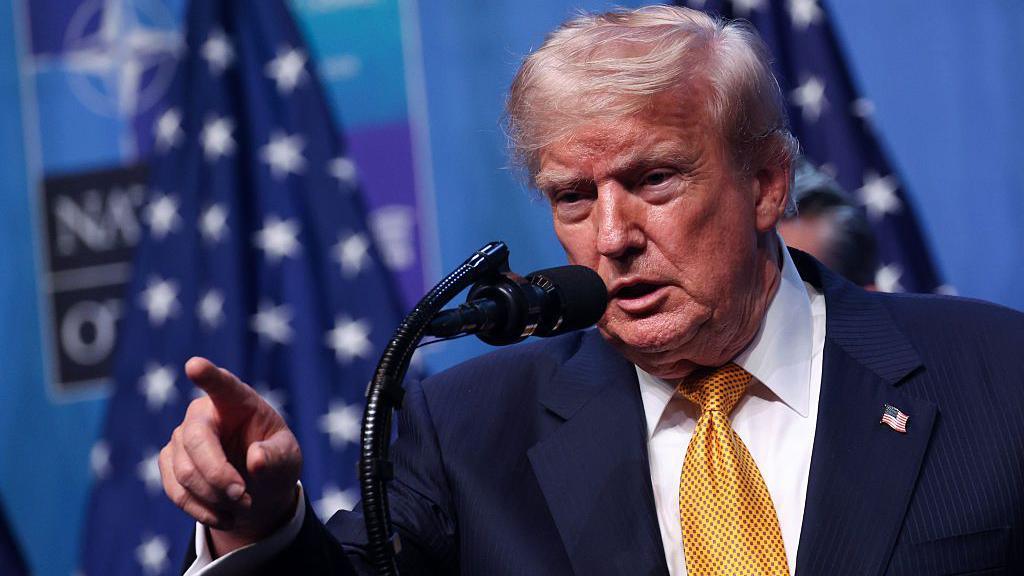 US President Donald Trump wearing a yellow tie, white shirt, blue suit and American flag pin. He stands at a microphone and is pointing, his mouth open.