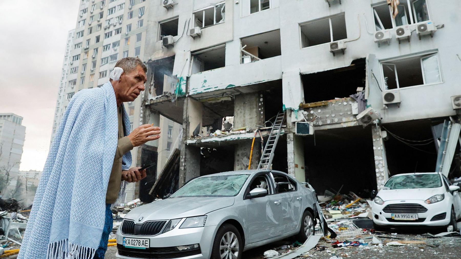 Injured man wearing a blue blanket standing in front of an apartment building that has been badly damaged