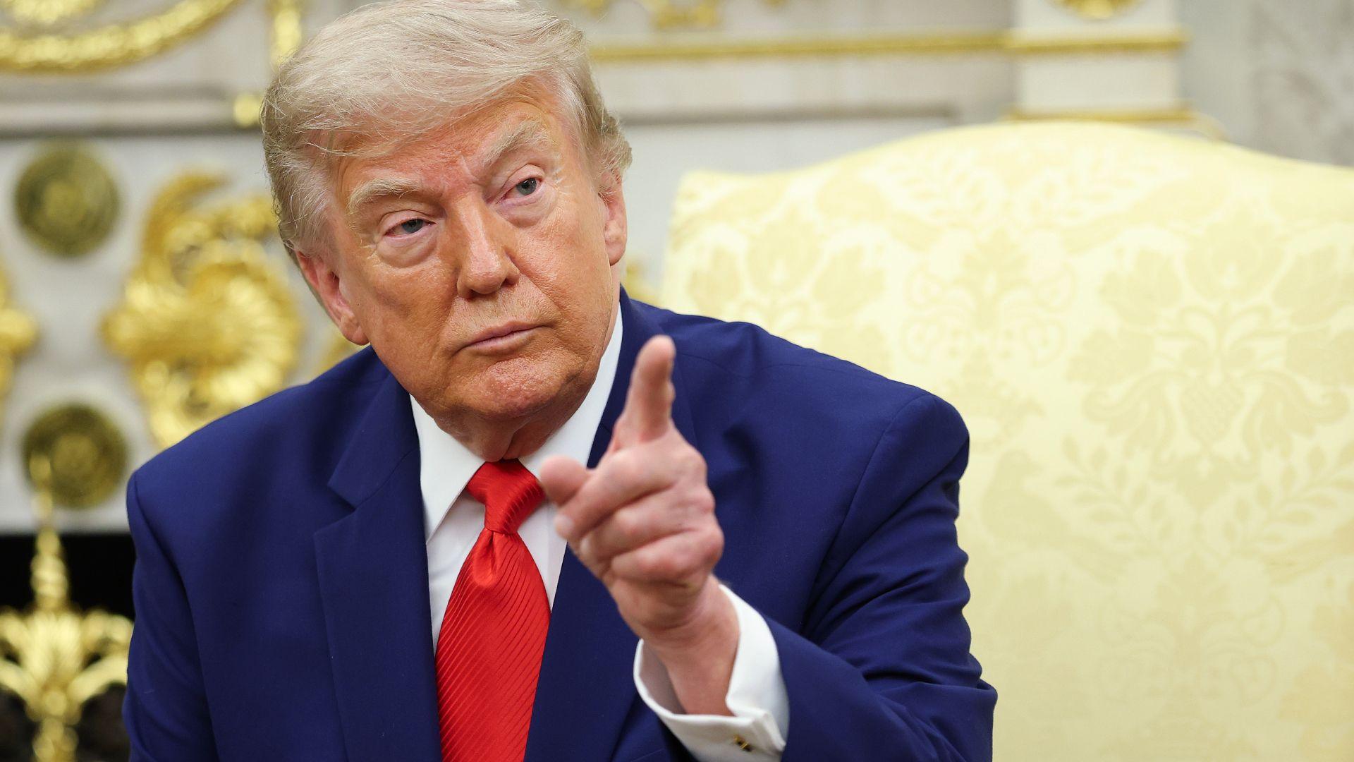 President Donald Trump pointing his finger. He has a serious look on his face and is wearing a blue suit with a white shirt and red tie.