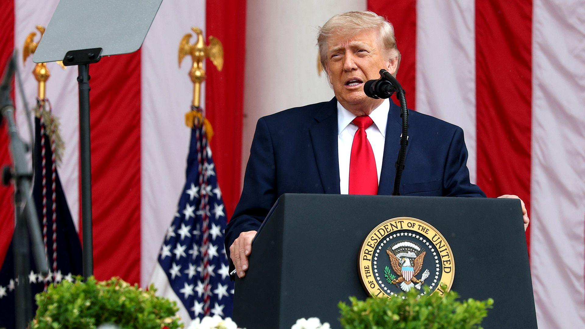 Donald Trump stands at a lecturn with a plaque reading ' President of the United States' with the US eagle logo, as he speaks into a microphone while wearing a dark suit and red tie against the backdrop of the US flag.
