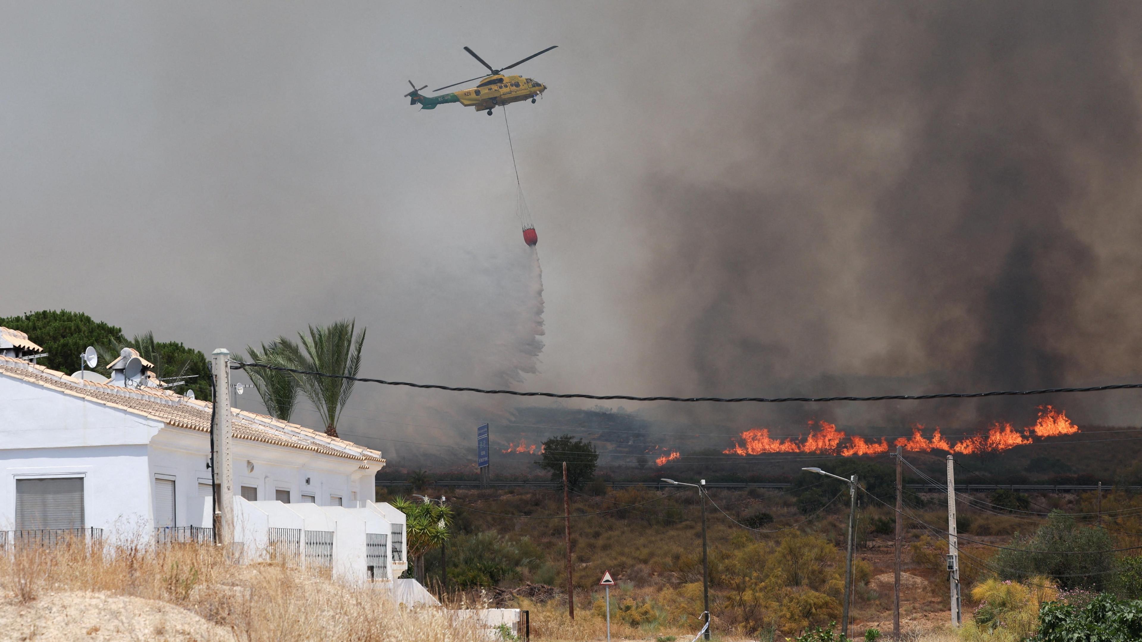 A helicopter with a large bucket attached drops water on flames with a house in the foreground
