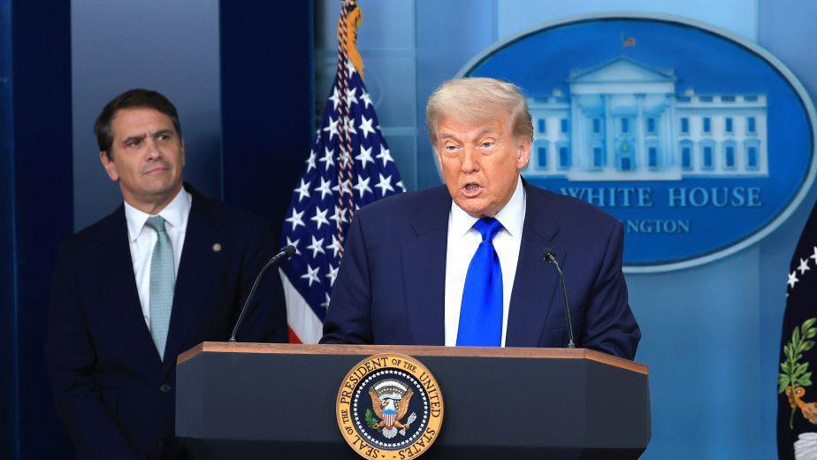 Trump is at the podium and speaking while wearing a dark blue suit and bright blue tie. To his right and behind him is Todd Blanche who is wearing a dark suit and indigo tie