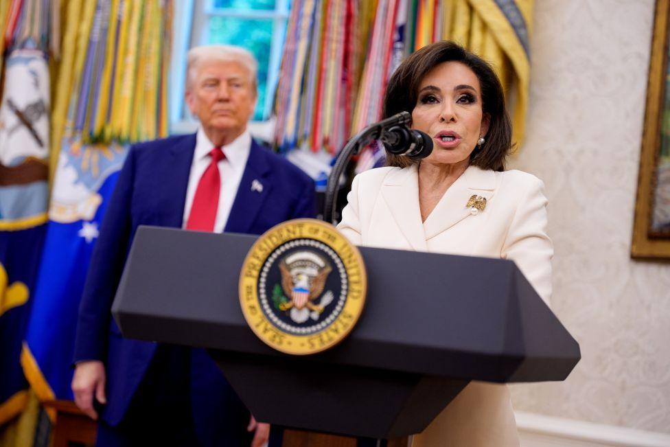 Jeanine Pirro is in the Oval Office speaking from a podium bearing the presidential crest. She is wearing a white stui and a gold brooch on her left lapel. Donald Trump is standing behind her, slightly out of focus, wearing his trademark royal blue suit and red tie.