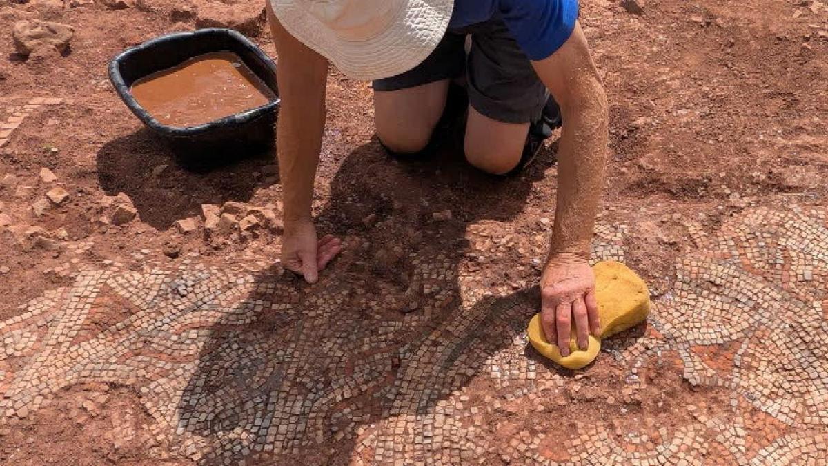 A man uses a yellow sponge to scrub a Roman mosaic found in a farmer's field in Devon. He is wearing a white sun hat and blue t-shirt.