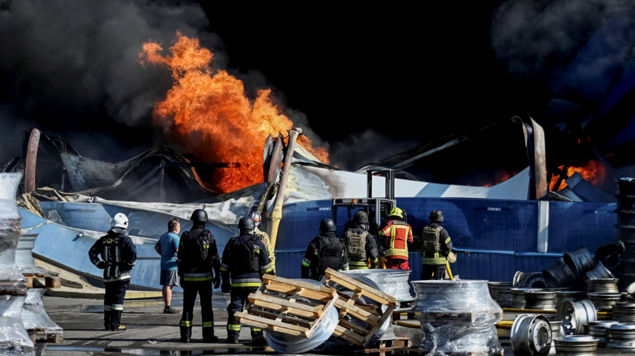 Wide shot showing Ukrainian firefighters in black and yellow uniforms observing flames rising from the debris of what appears to be a warehouse following a drone strike.