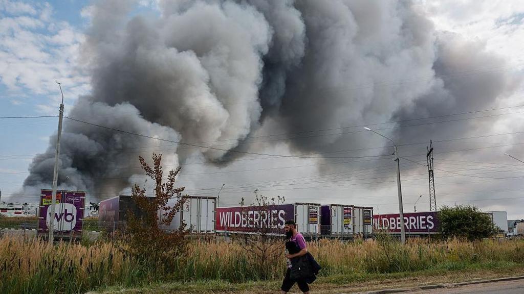 Wildberries employee walks past plumes of smoke rising above wildberries logistics complex. There are seven company lorries in front of the smoke as well as roadside shrubs