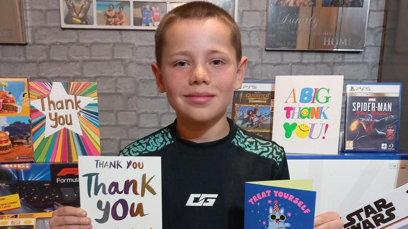 Phoenix is holding two thank you cards and is surrounded by gifts. He has short brown hair and is smiling.