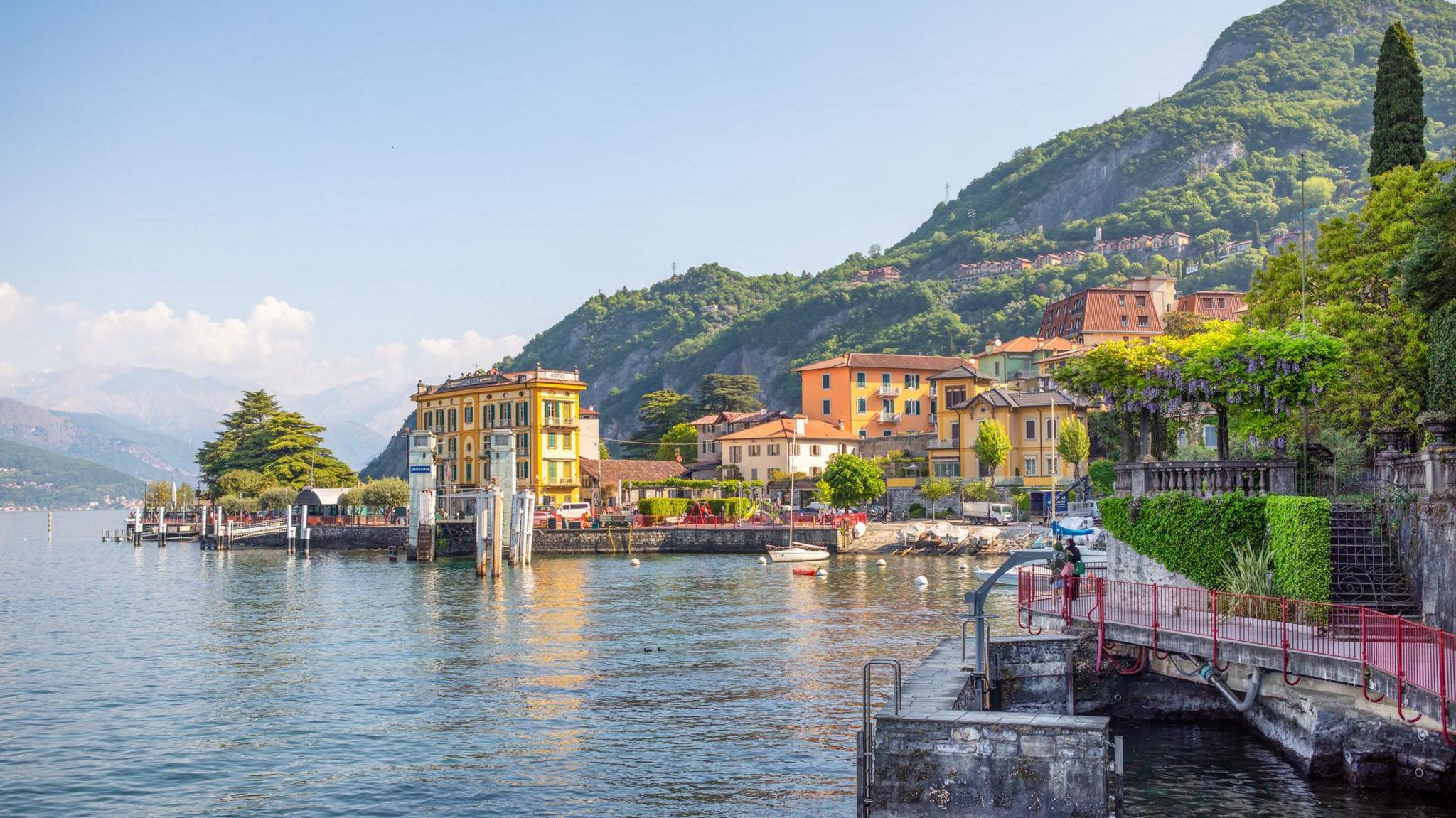 Sunny yellow buildings and greenery sit infront of green mountains overlooking Lake Como.