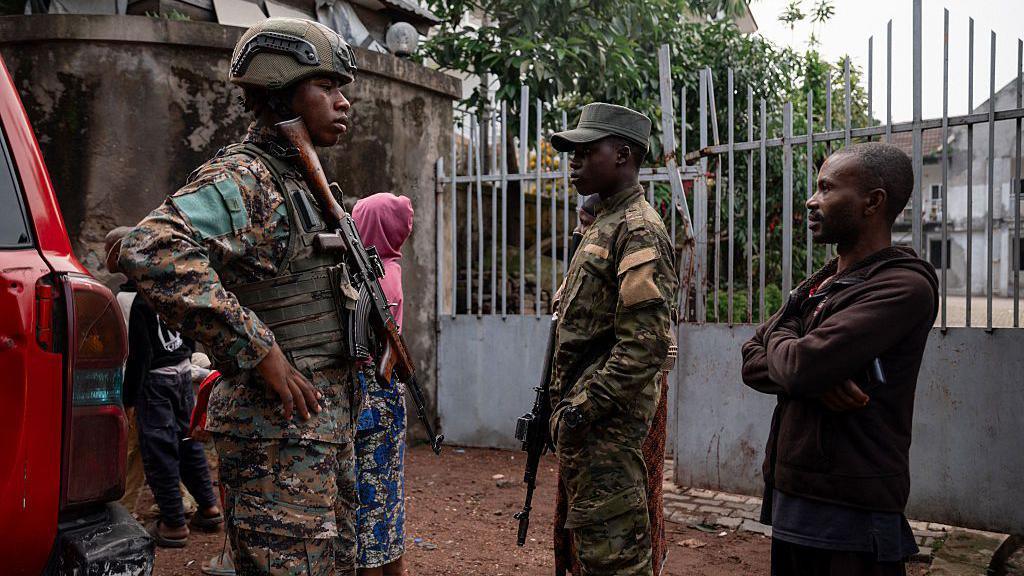 Two men dressed in military uniform and holding rifles stand alongside a member of the public in a brown hoody and trousers. A woman in a pink hoody has her back to the camera in the background