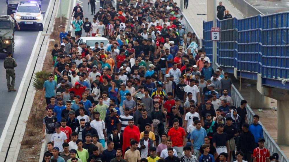 A large group of migrants. mainly men, being escorted along a walkway by police.