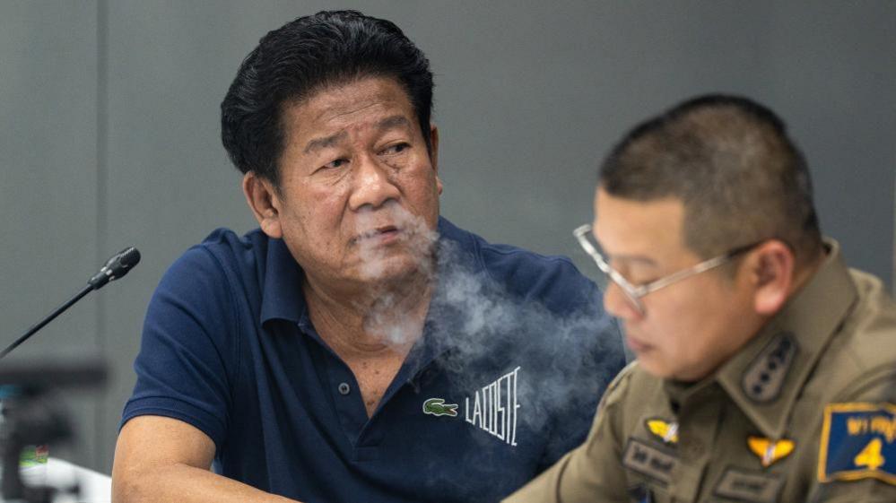 Former lawmaker Chalong Riewrang in a blue t-shirt exhales out cigarette smoke at a police station where he was allowed to hold a press conference after allegedly shooting and killing former police officer Thongchai Yenprasert. A police officer in uniform and glasses is seated to his right.