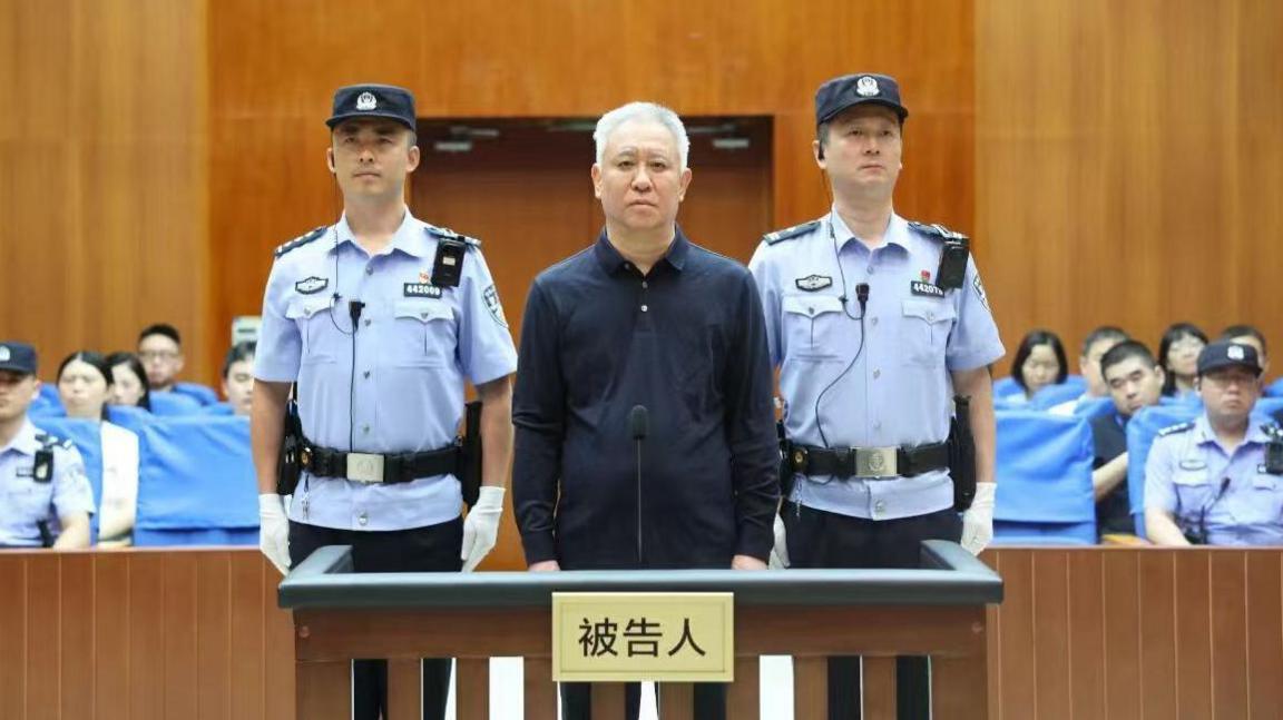 China Evergrande Group founder Hui Ka Yan takes the stand in a court in China. He has a blank look and is standing between two officers, with members of the audience in the background.